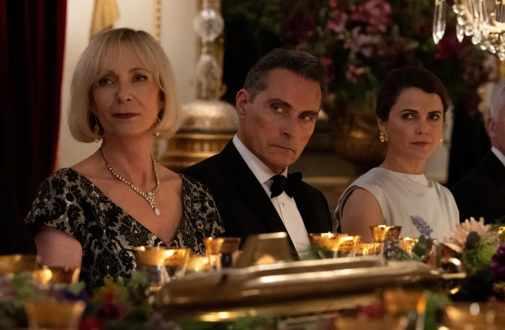 Allison Janney, in a black gown, Rufus Sewell, in a black tuxedo, and Keri Russell, in a silver gown, seated at a formal dinner table.