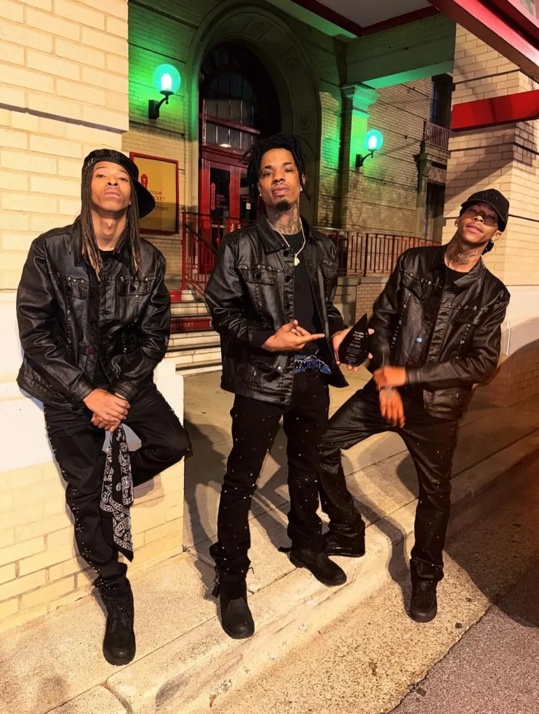 Three men in black outfits with studded jackets pose in front of a brick building.