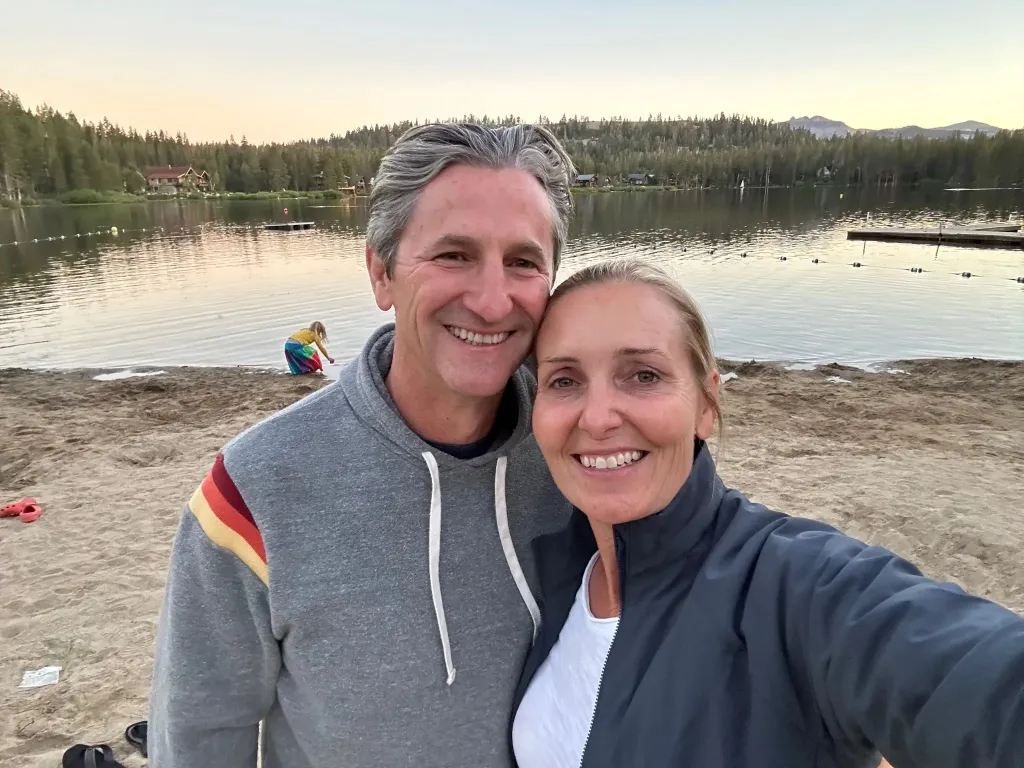 Amy Hillyard and her husband, Chris Hillyard, smile for a photo with a lake and forest in the background.