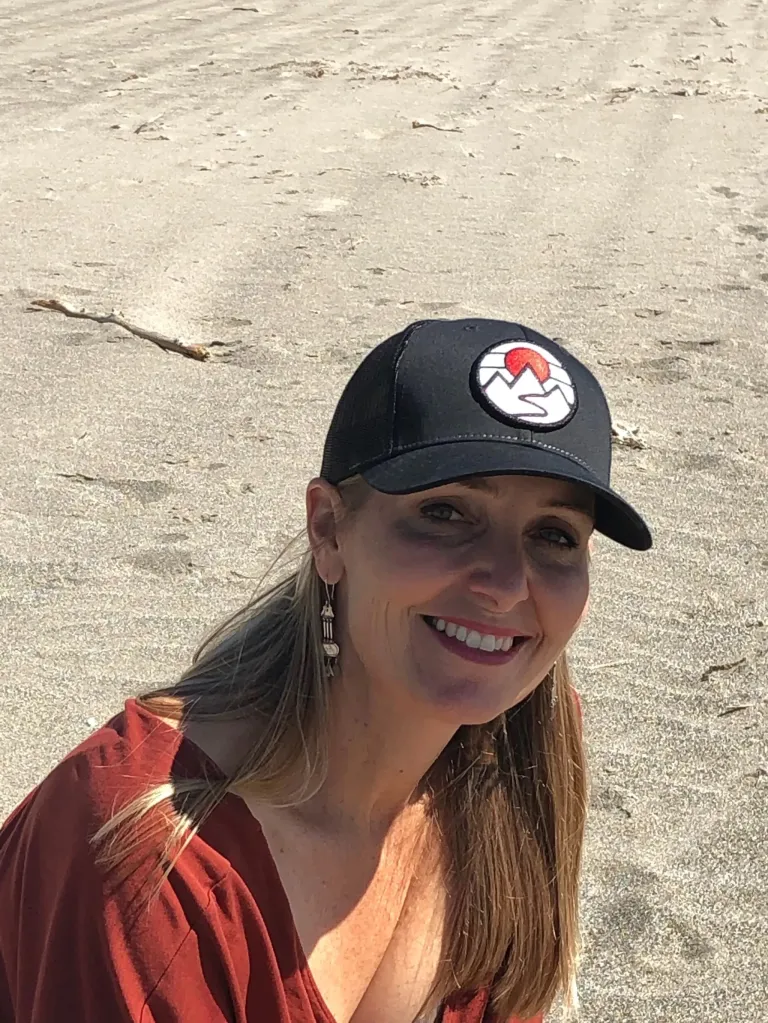 Amy Hillyard, with long blonde hair, smiles at the camera while wearing a black baseball cap and a rust-colored top.