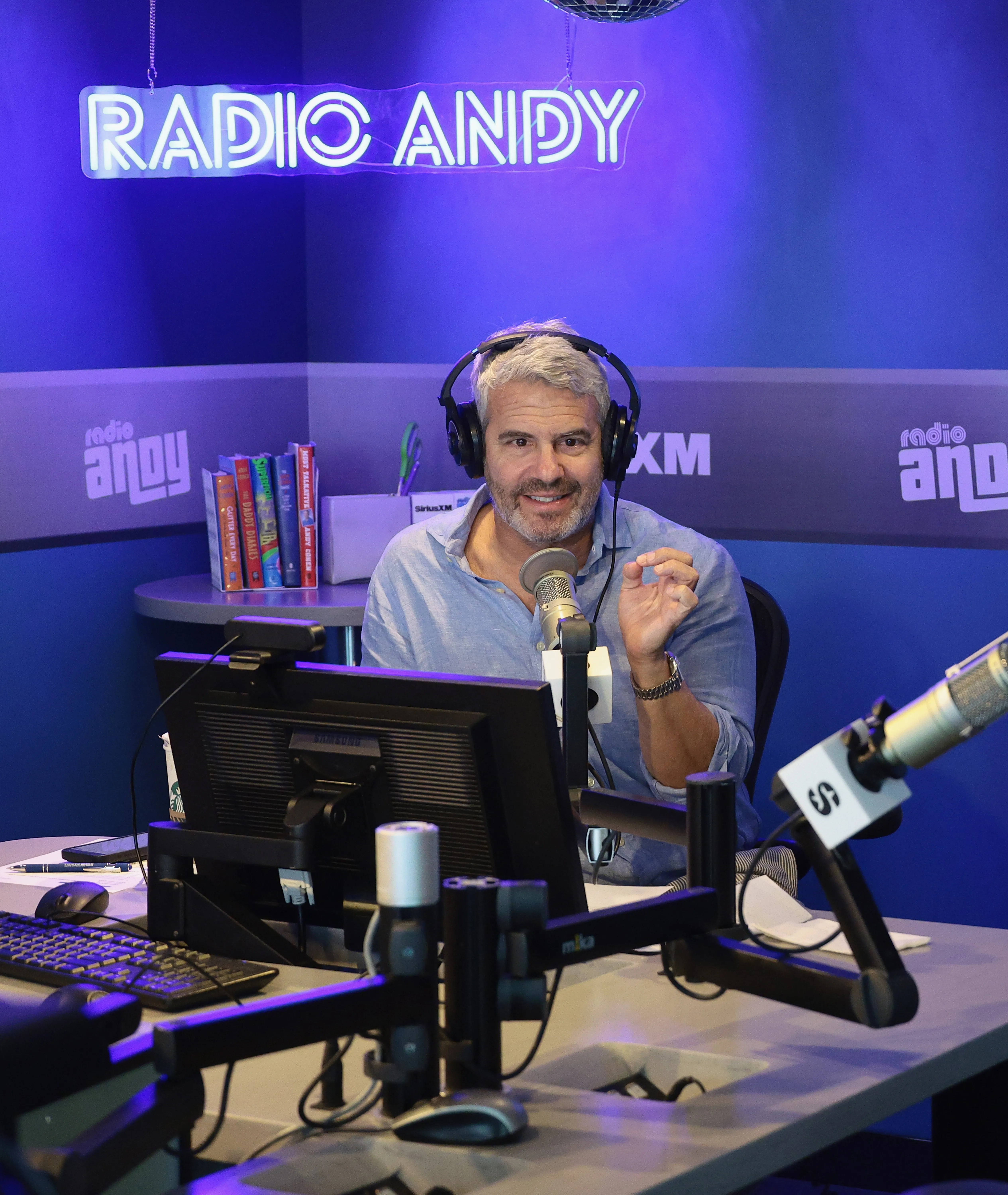Andy Cohen sitting in a radio studio with headphones on, speaking into a microphone, with a 