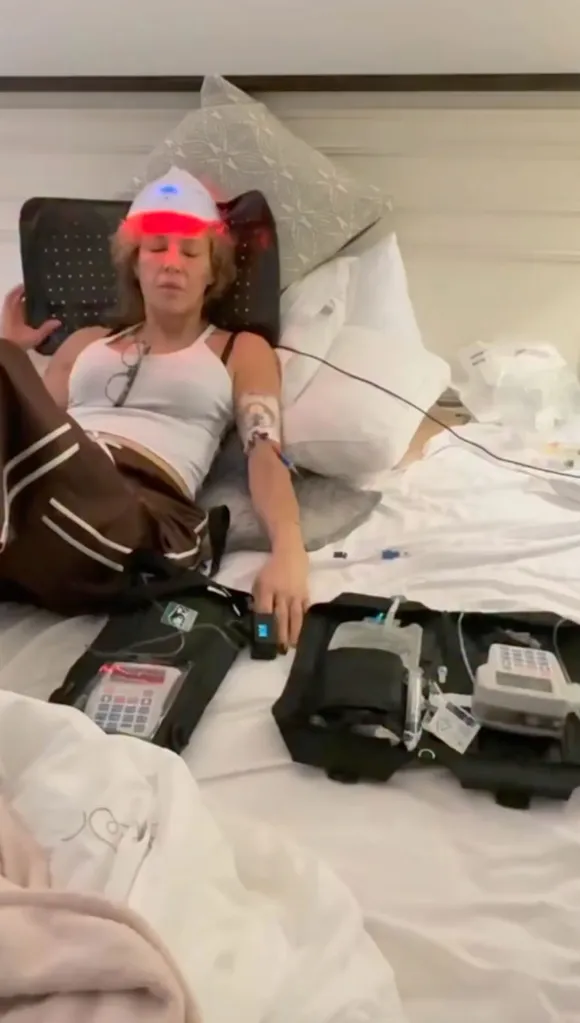 Woman lying in bed with an IV drip in her arm, a light therapy mask on her head, and medical equipment beside her.