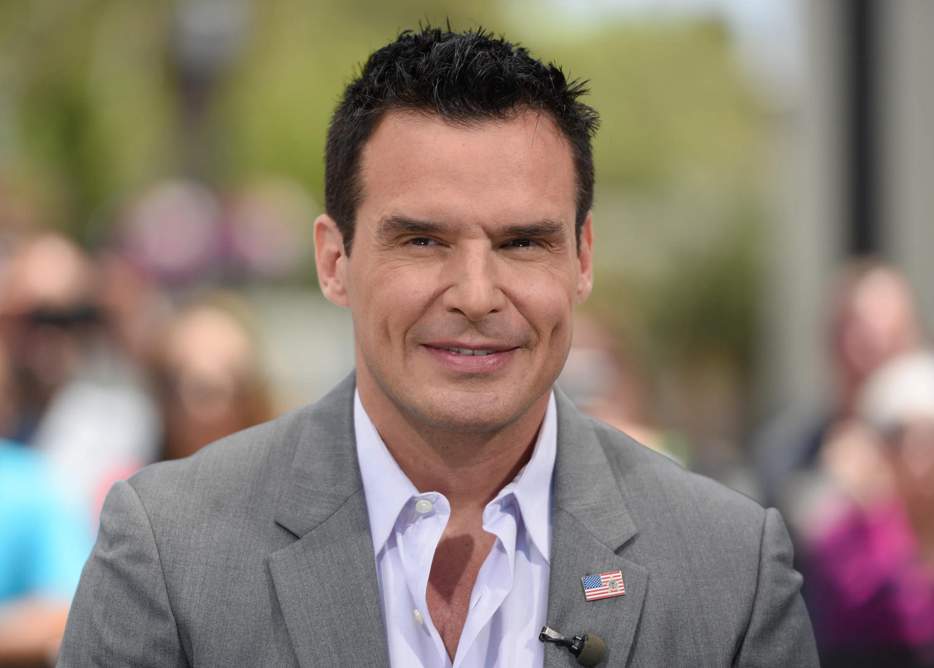 Antonio Sabato Jr. smiles while wearing a light grey suit jacket with an American flag lapel pin and a white button-down shirt.