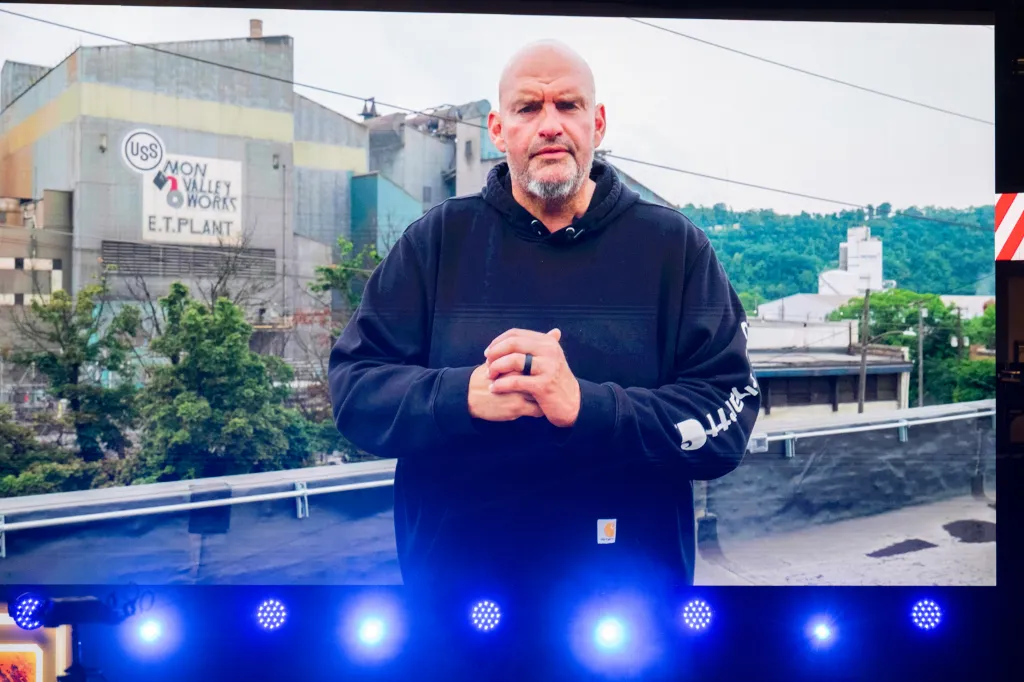 John Fetterman appearing by video on a large screen with an industrial building in the background.