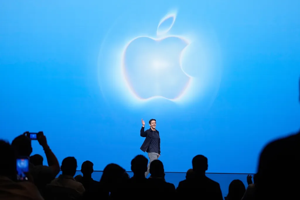 John Ternus waves on stage in front of a blue screen with a glowing Apple logo.