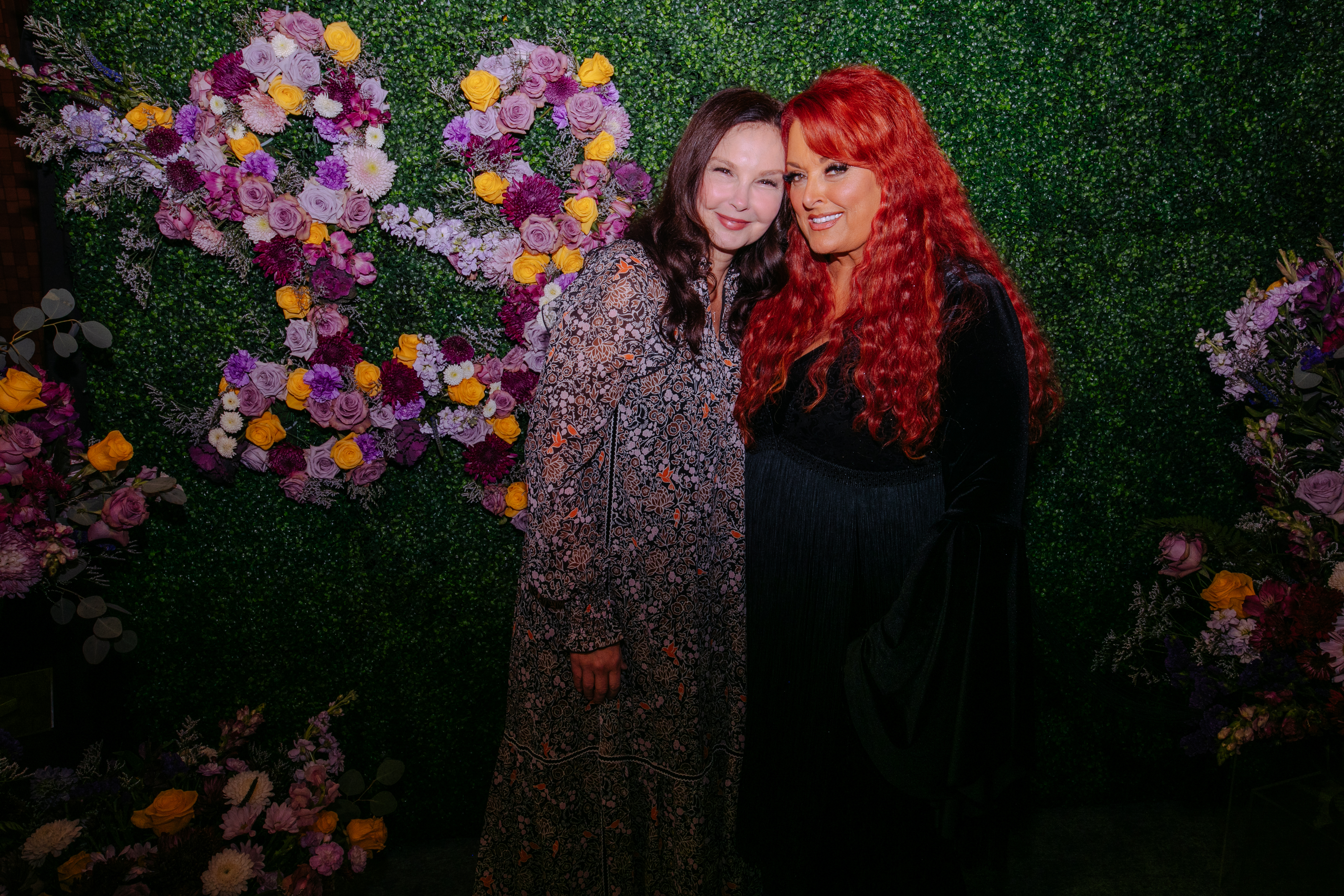 Ashley Judd and Wynonna Judd posing together against a green wall decorated with flowers.