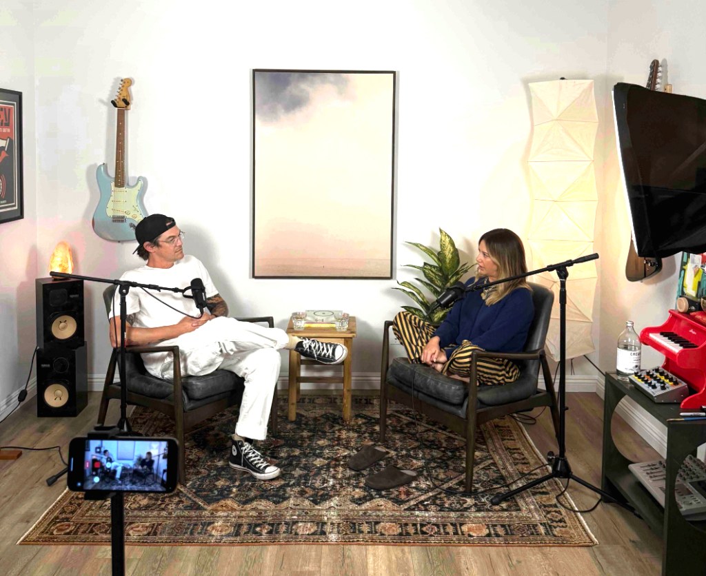 Christopher French and Ashley Tisdale sit in chairs facing each other in a recording studio, with microphones and a smartphone on a tripod in the foreground.