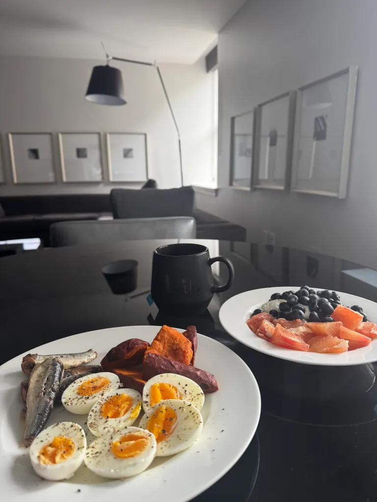A black mug sits on a glossy table next to two plates of food: one with sardines, halved boiled eggs, and sweet potatoes; the other with grapefruit, blueberries, and a dollop of cream.