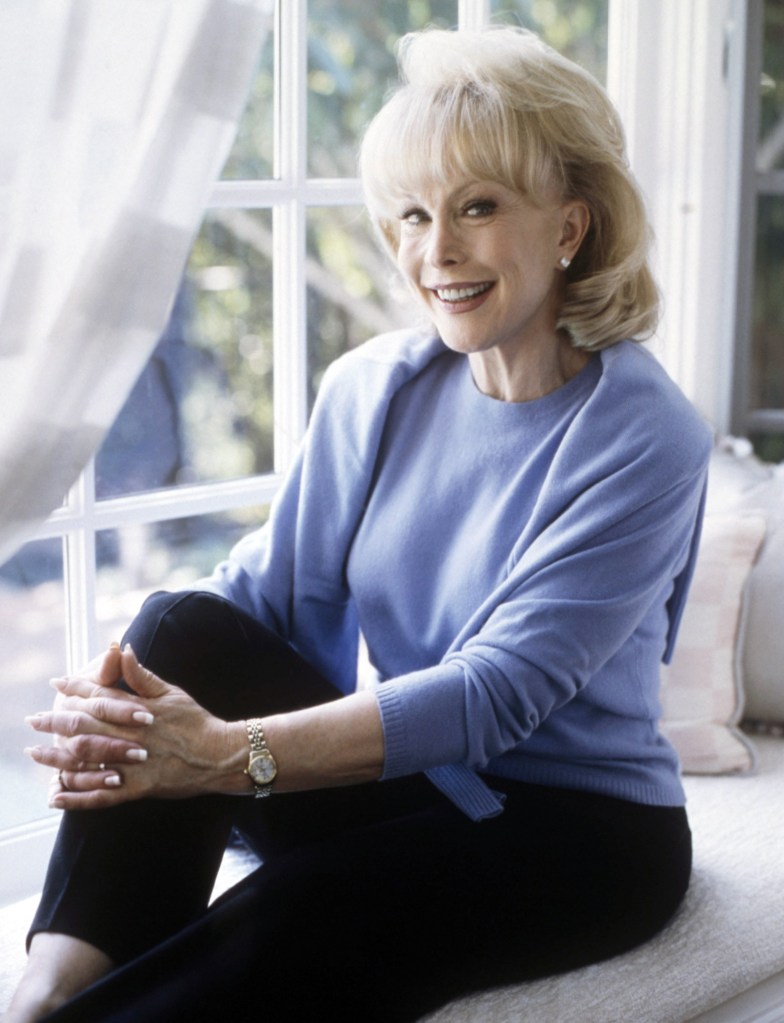 Barbara Eden in a blue sweater set sitting on a window seat.
