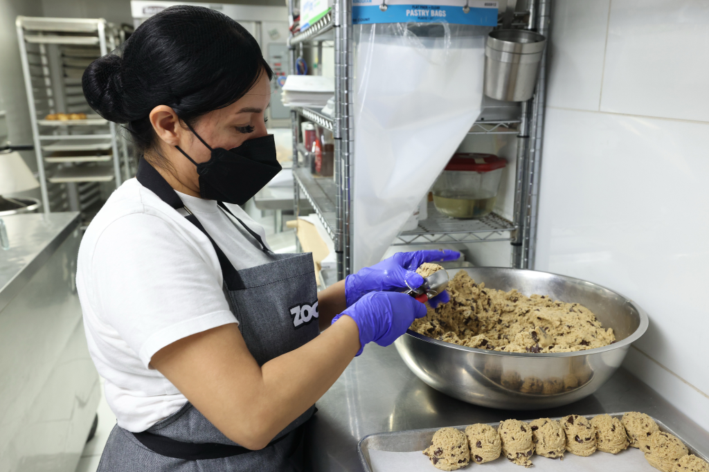 A baker in a black face mask and purple gloves portions chocolate chip cookie dough from a large silver bowl onto a baking sheet.