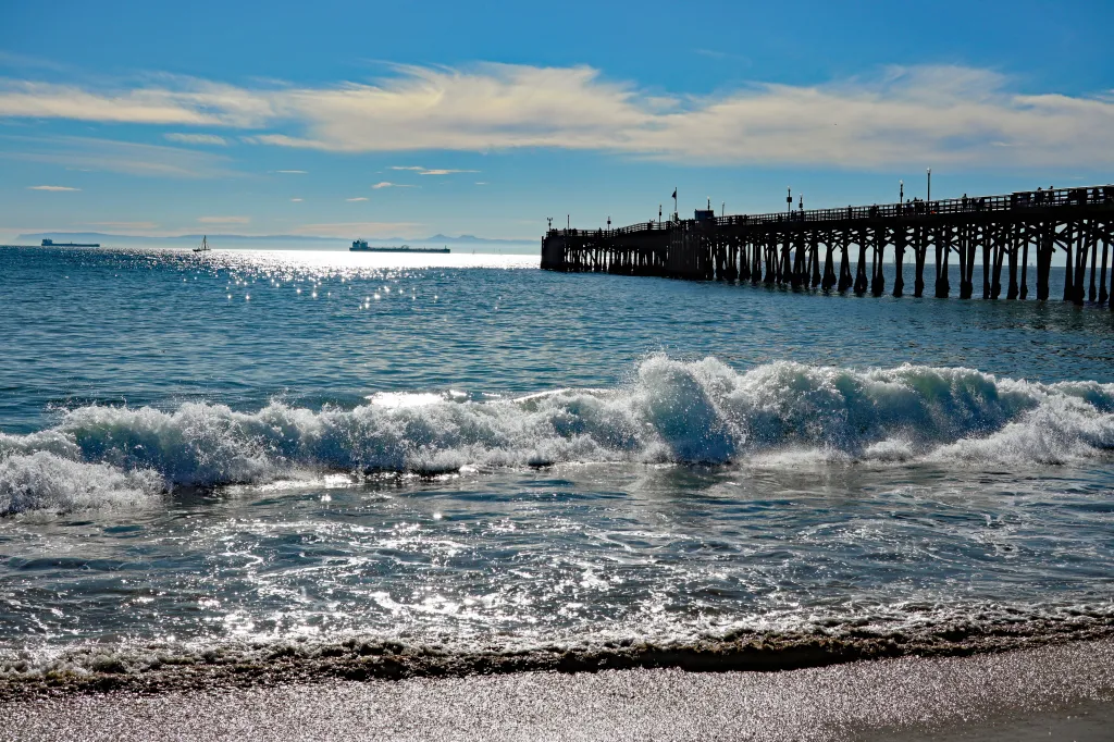 Waves crash along the sands of Seal Beach, California, with sunshine reflections, a city pier, ships, sailboats, and Catalina Island on the distant horizon.