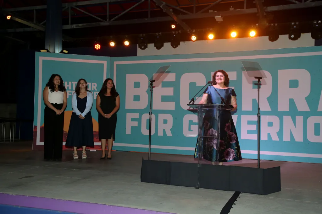 Dr. Carolina Reyes speaks at a podium with her daughters Clarisa, Natalia, and Olivia standing nearby during the Xavier Becerra for Governor election night party.