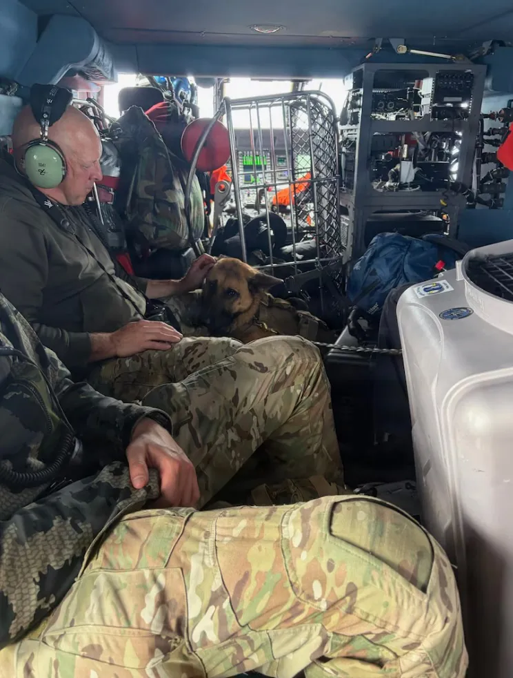A man in a military helicopter petting a German Shepherd dog.