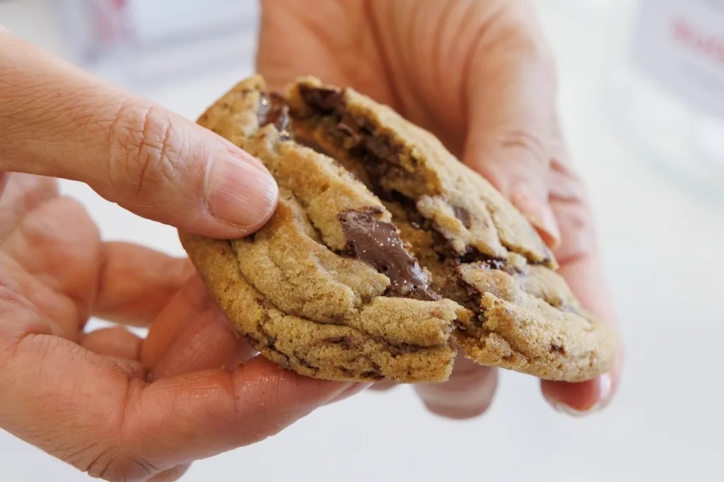 A hand breaks apart a gooey chocolate chip cookie, revealing melted chocolate inside.