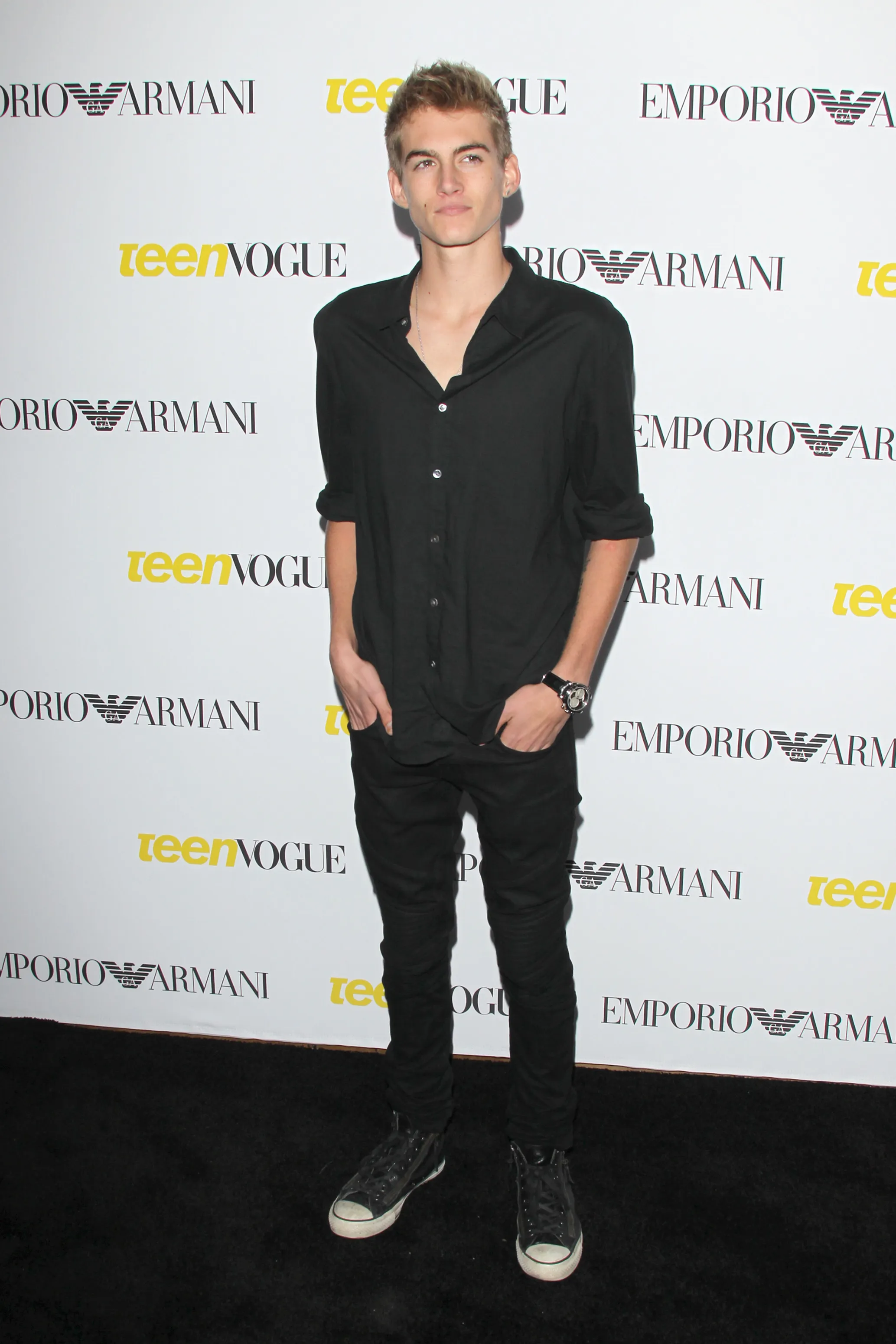 Full-length portrait of Presley Walker Gerber at an event, wearing a black shirt and pants with black sneakers, posing in front of a white backdrop with 