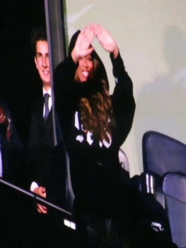 Beyoncé smiles and covers her face with her hands, wearing a black hoodie with white letters, while a man in a suit smiles behind her.