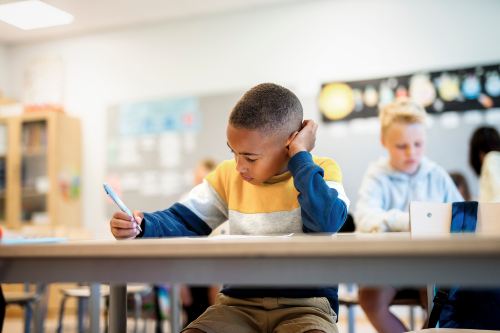 A boy writing on paper while sitting at a desk in an elementary school classroom.
