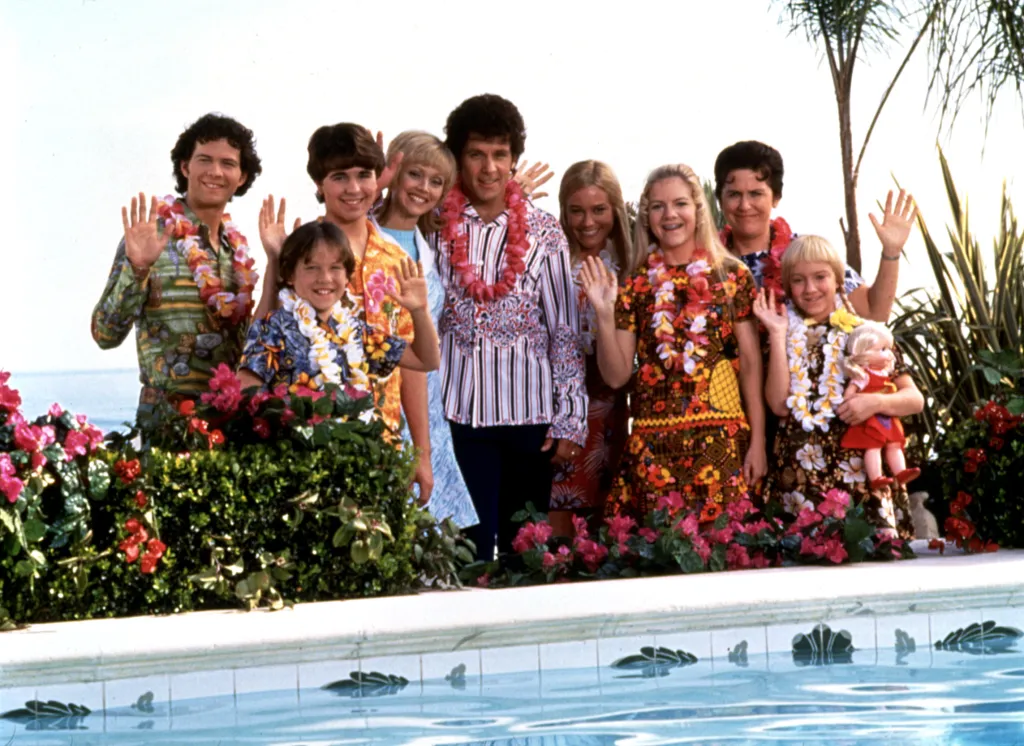 A group of 10 people, including children and adults, wearing leis and Hawaiian shirts, waving at the camera from a poolside setting, with the ocean in the background.