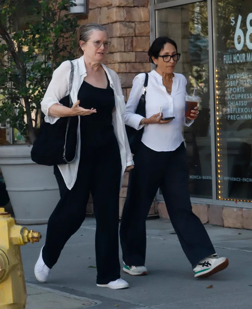 Bridget Fonda wearing a white blouse, black top, black trousers, and white trainers, walking alongside another woman.