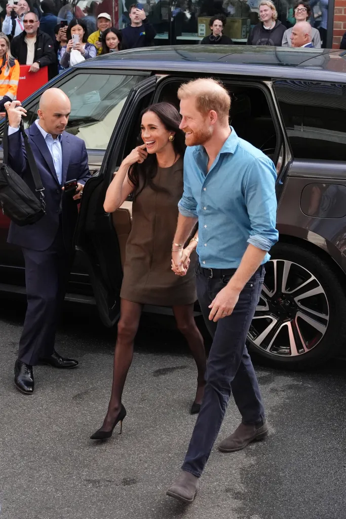 Prince Harry and Meghan, Duchess of Sussex, holding hands and smiling as they exit a car to a crowd of onlookers.