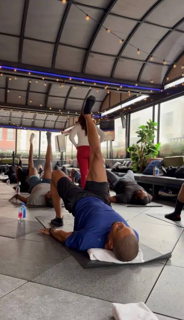 Men participate in a Bro-lates Pilates class, lying on mats with legs raised, led by a female instructor.