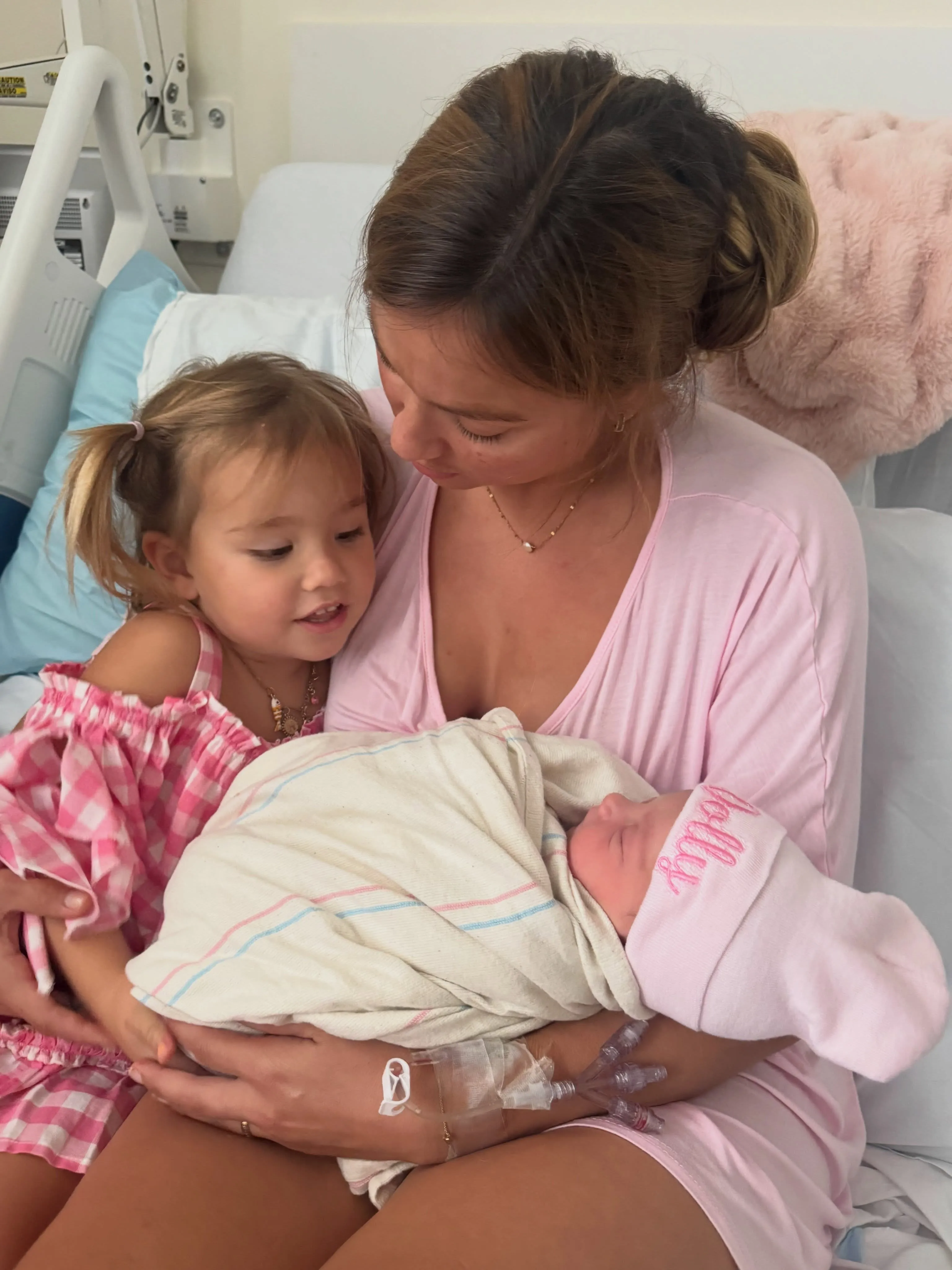 A mother wearing a pink shirt holds her newborn baby wrapped in a blanket, with her older daughter in a pink and white checkered dress looking at the baby.