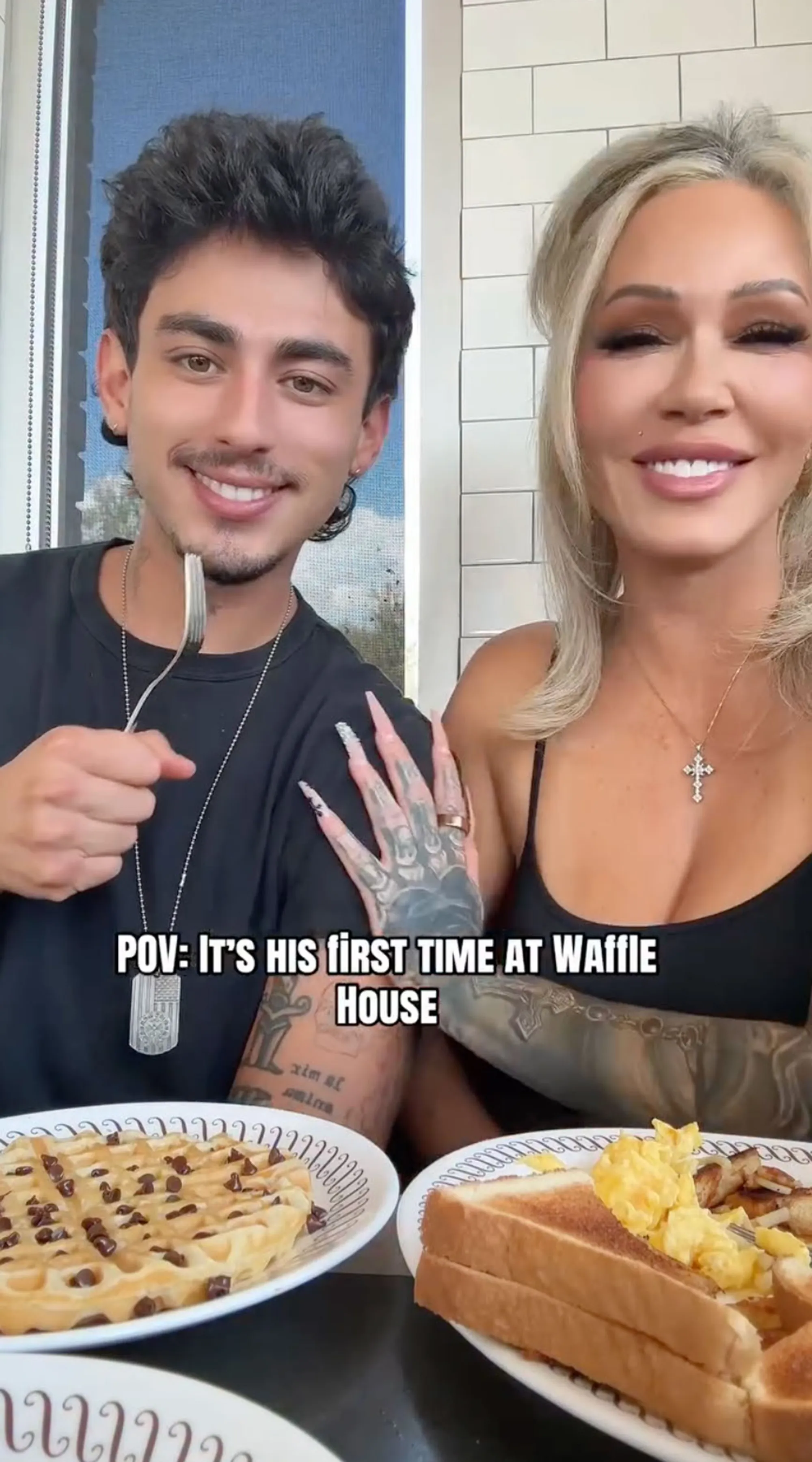 A man with dark hair holding a fork, smiling at the camera, next to a blonde woman smiling, with plates of breakfast food in front of them.