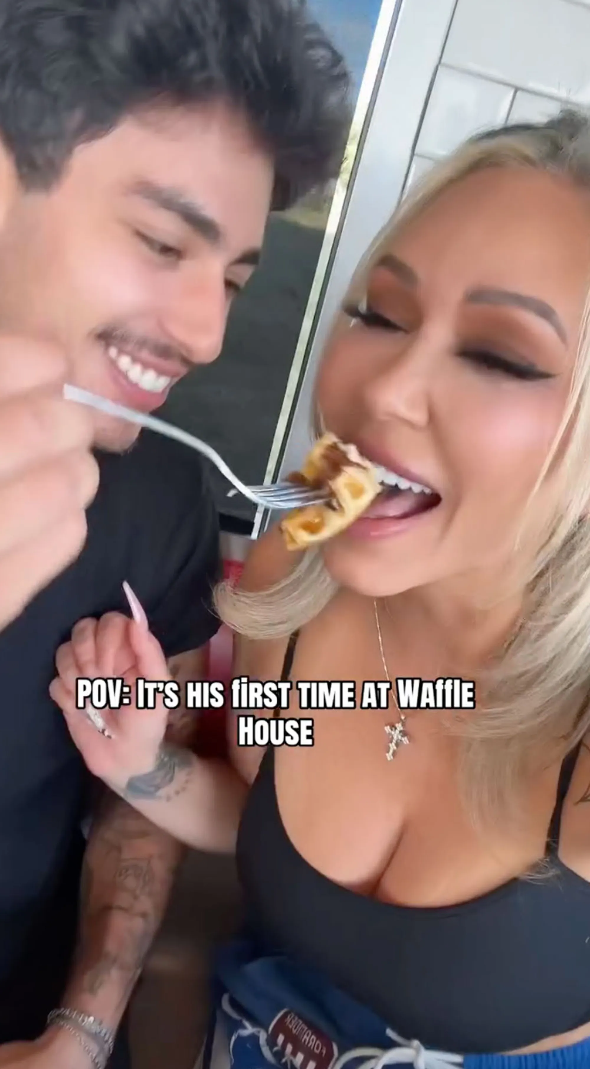 A man with a mustache smiles while feeding a woman a piece of waffle with a fork.