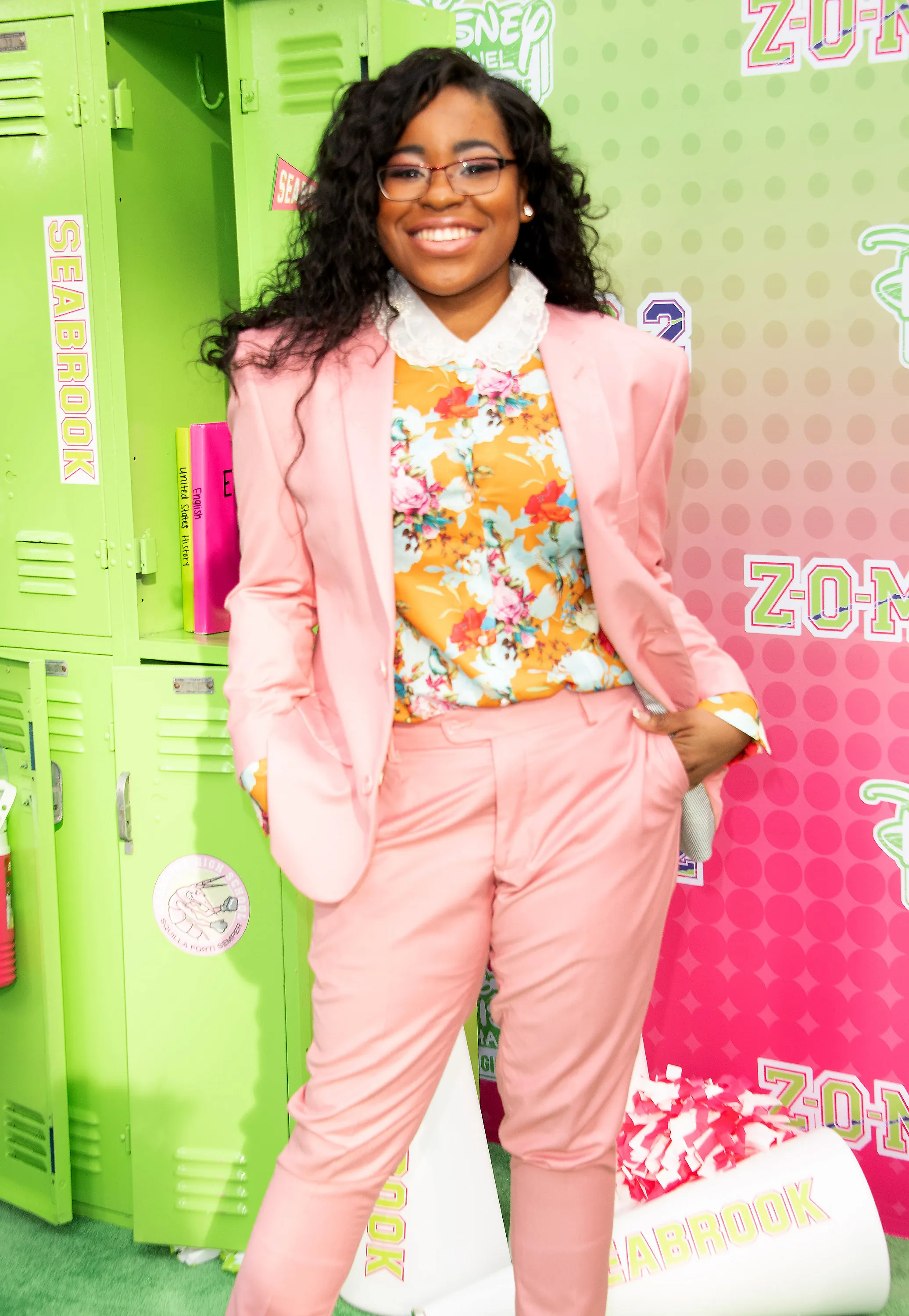 Carla Jeffery smiling in a light pink pantsuit, floral shirt, and glasses, standing in front of lime green lockers.