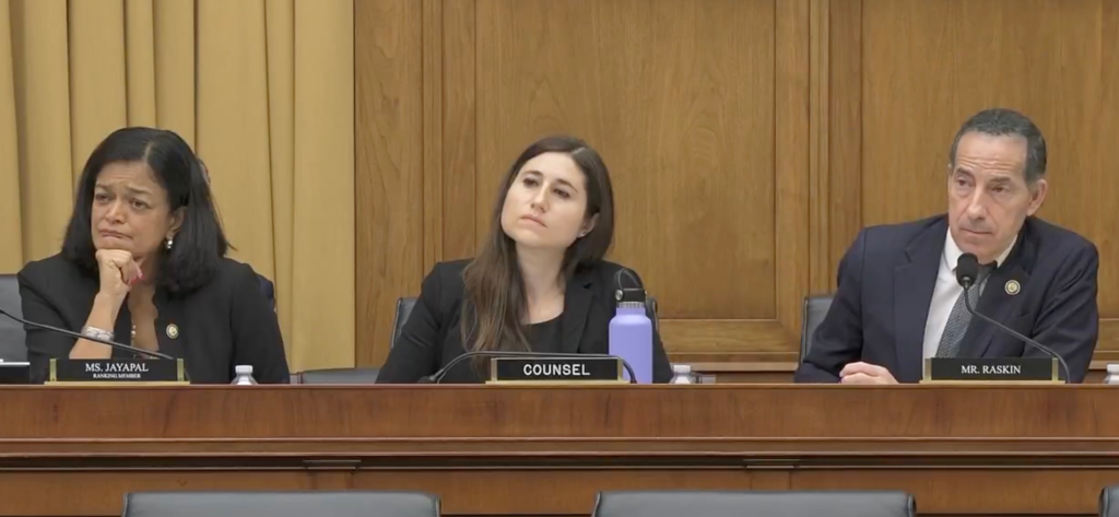 A dark-haired woman, a brunette woman, and a white man seated at a long wooden desk.