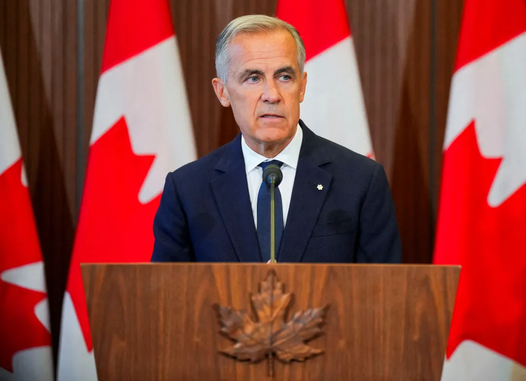 Canada's Prime Minister Mark Carney speaking at a podium with Canadian flags in the background.