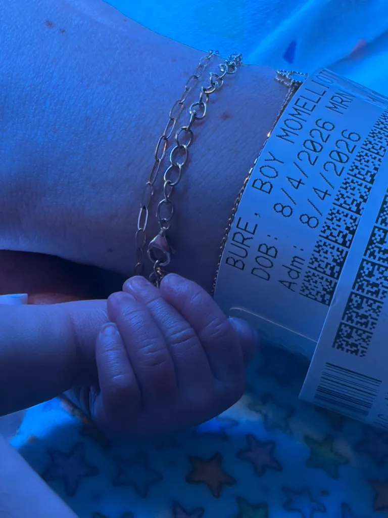 A baby's hand clutching an adult's finger, illuminated in blue light, with a hospital band visible on the adult's wrist.