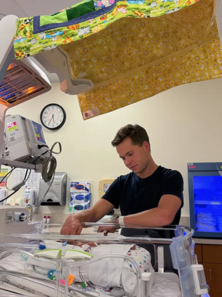 A man looking at a newborn baby in an incubator, with a clock on the wall and medical equipment nearby.