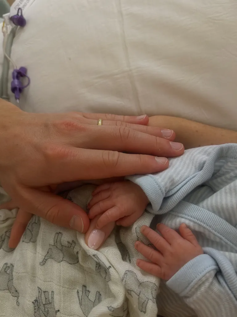 An adult's hand, wearing a thin gold band on the ring finger, holds the hand of a newborn baby.