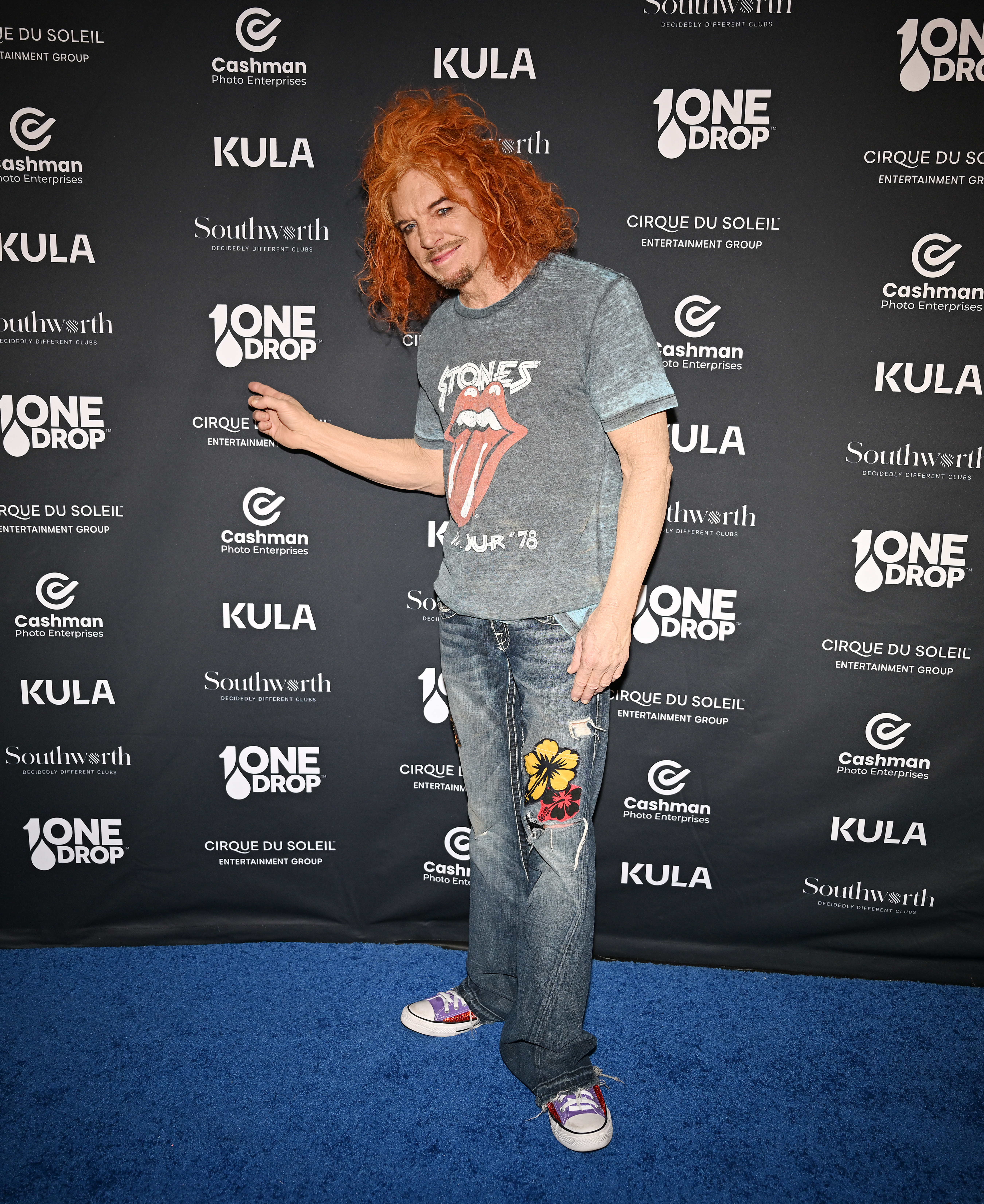 Carrot Top, wearing a Rolling Stones t-shirt, stands on a blue carpet in front of a backdrop with event logos.