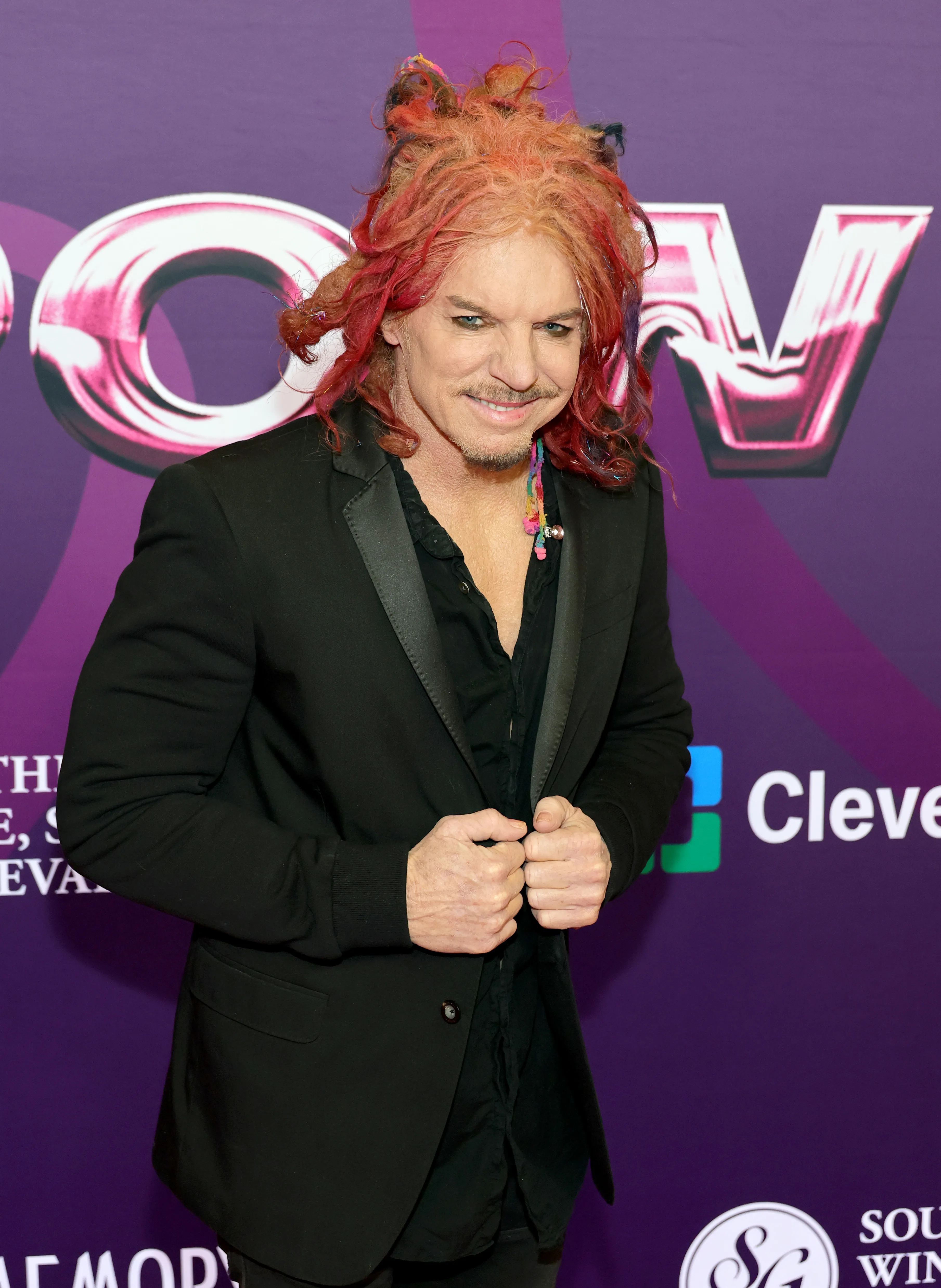 Comedian Carrot Top with red, spiky hair, mustache, and goatee, wearing a black blazer and shirt, stands in front of a purple background with white and pink lettering.