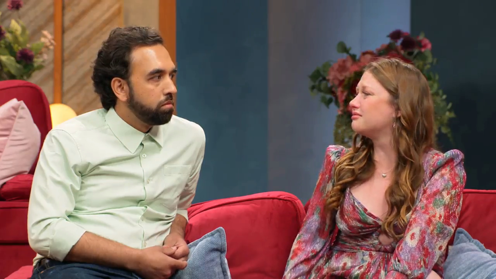 Catie looks away from Josh as she cries, sitting on a red couch.
