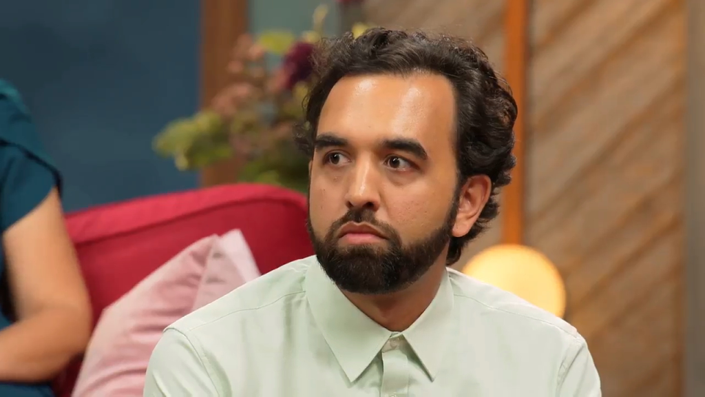 A man with dark curly hair and a beard looks to the left, wearing a light green button-up shirt.