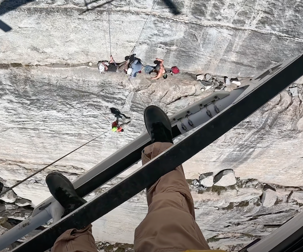 View from a helicopter of an injured rock climber being rescued from a steep cliff face by a rescue technician.