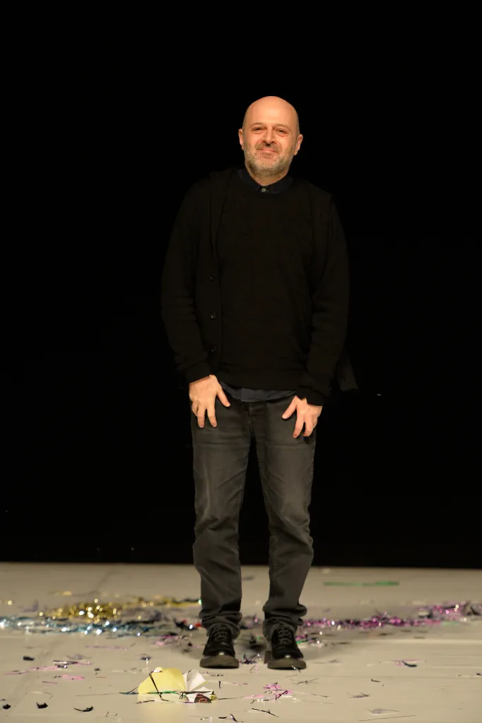 Hussein Chalayan on the runway, smiling and wearing a black cardigan and dark jeans.