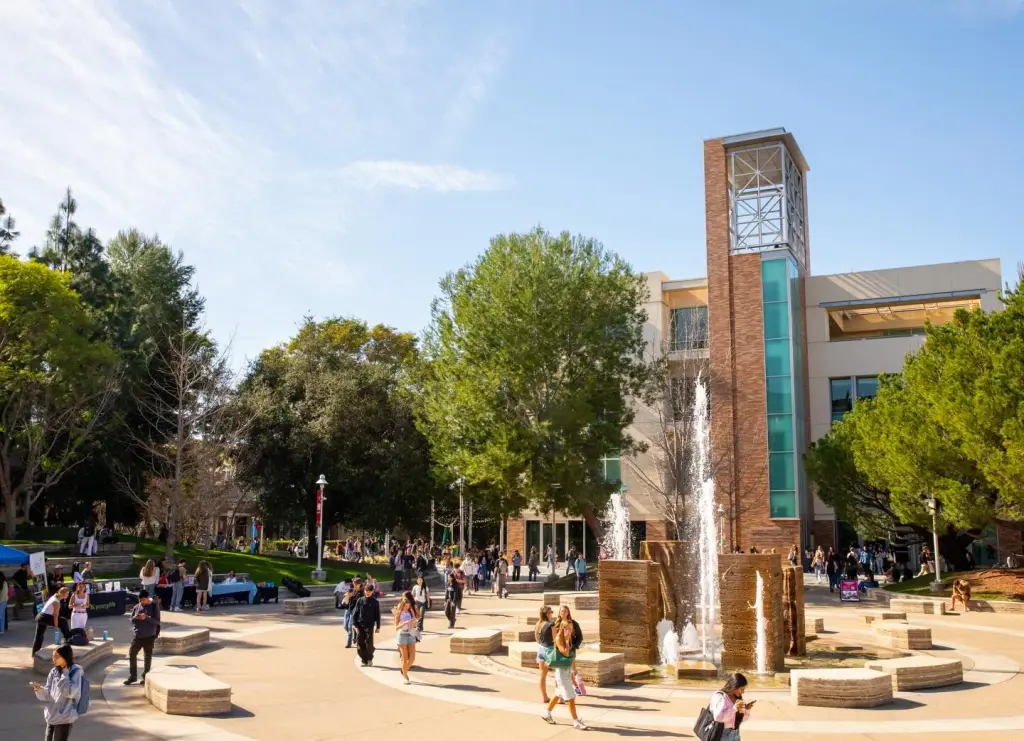 Students walk through the sunny campus of Chapman University, with a large fountain in the foreground and a modern brick building behind it.