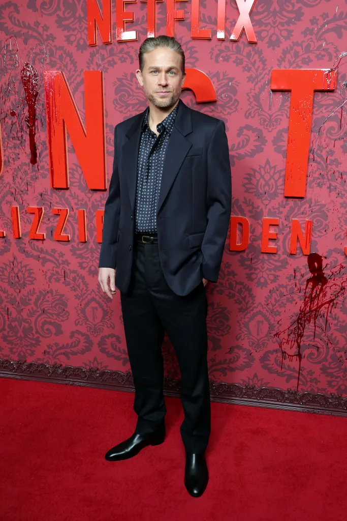 Charlie Hunnam poses on a red carpet in front of a red patterned backdrop for Netflix's 