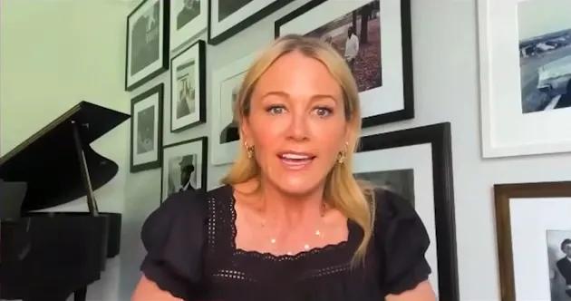 Christine Taylor speaks into the camera, with a black piano and framed pictures on the wall behind her.