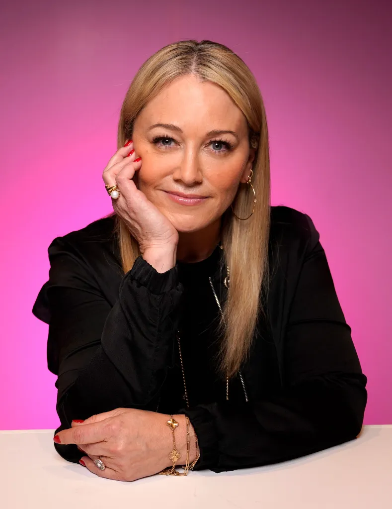 Christine Taylor smiling, resting her chin on her hand, against a pink background.