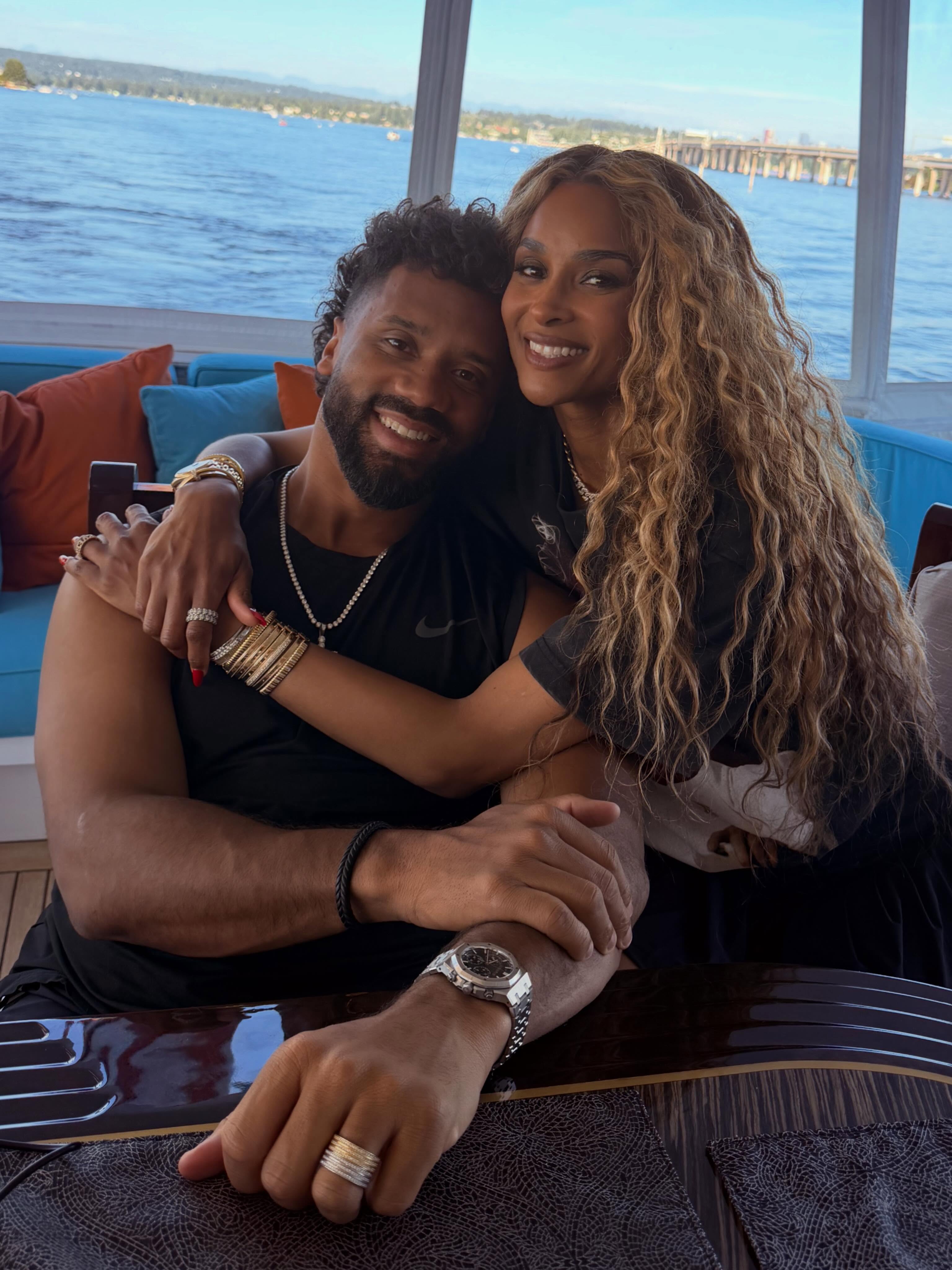 Ciara and Russell Wilson smiling and embracing on a boat with a body of water in the background.