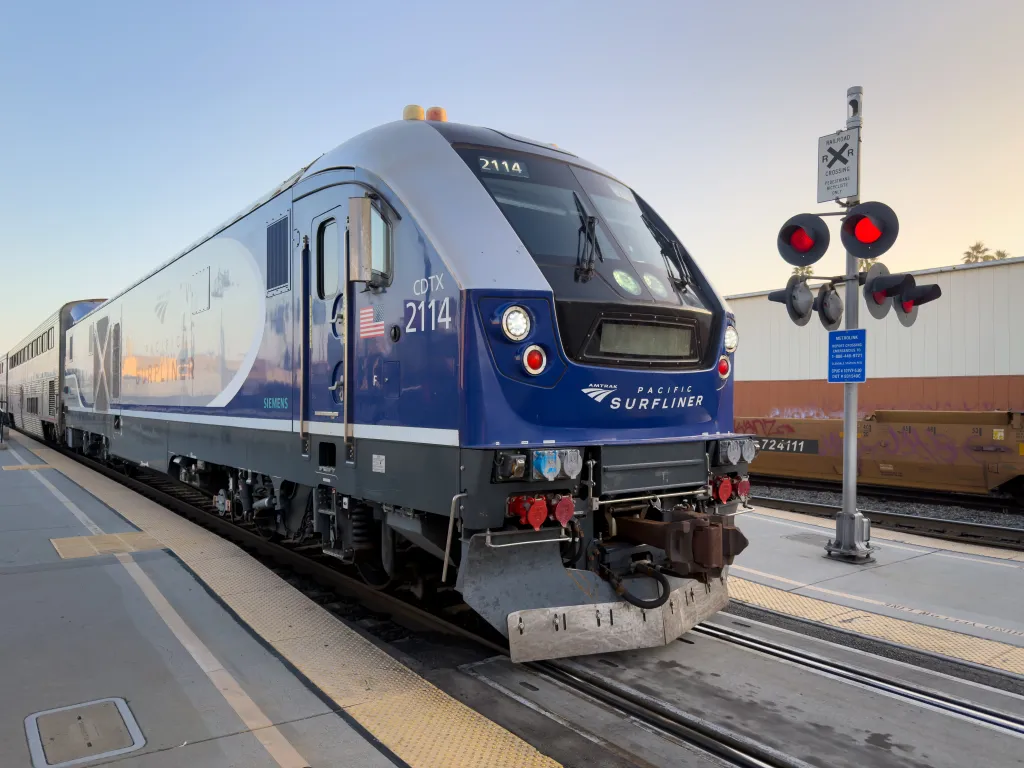 Close-up of an Amtrak Pacific Surfliner train locomotive at dusk, showing red crossing signals, bold blue livery, and Siemens branding.