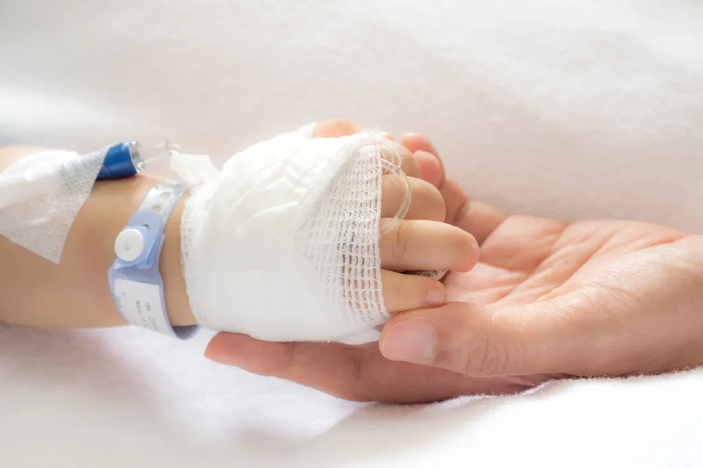 A child's bandaged hand with an IV drip and hospital wristband is gently held by an adult's hand.