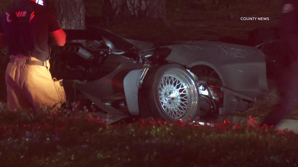 A dark-colored McLaren sports car split in half after a crash, with emergency personnel on the scene.