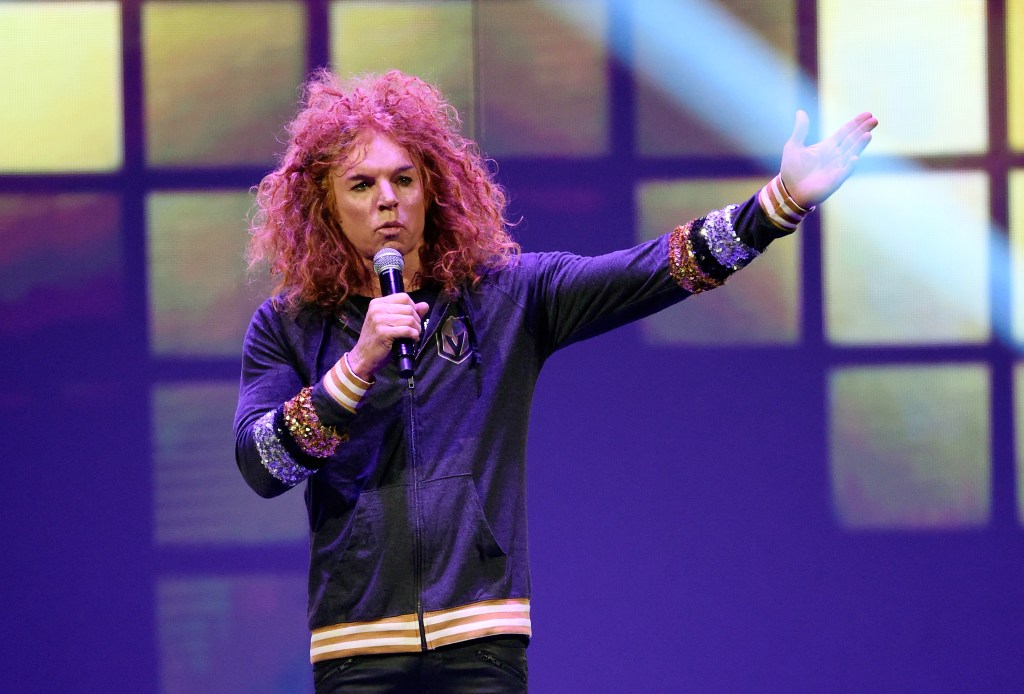 Comedian Carrot Top performing on stage, wearing a gray hoodie with sparkling sequined cuffs, holding a microphone in his left hand, and extending his right arm outwards.
