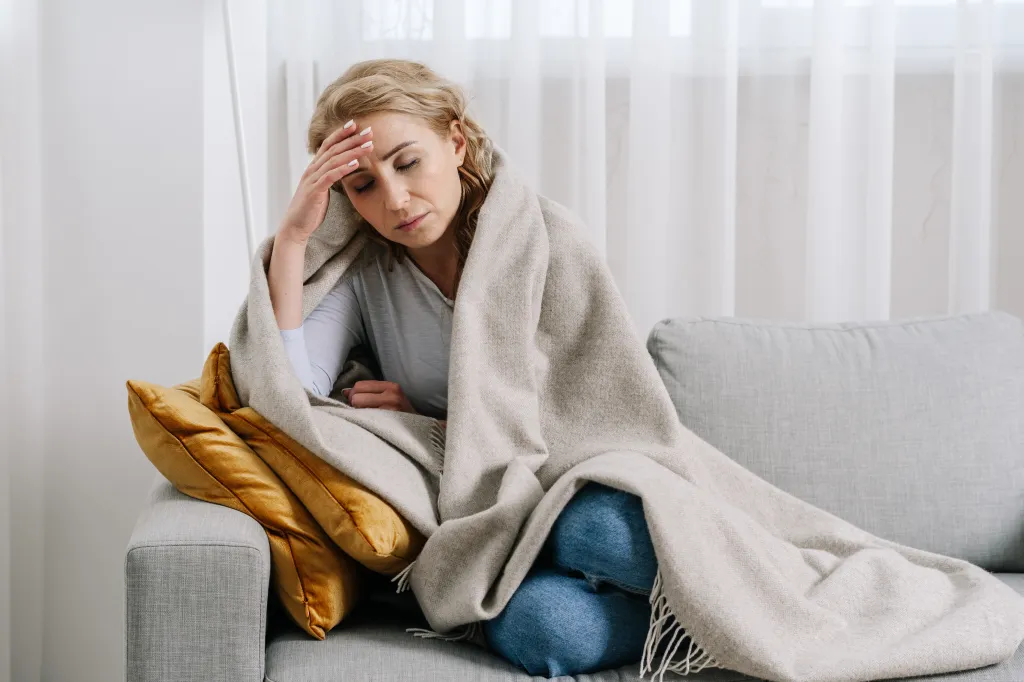 A sick woman covered with a blanket sits on a sofa, touching her forehead.